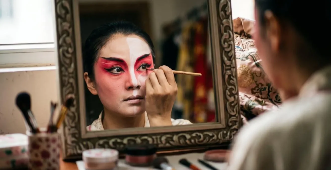Close-up of Cantonese opera artist applying intricate face paint in dressing room mirror